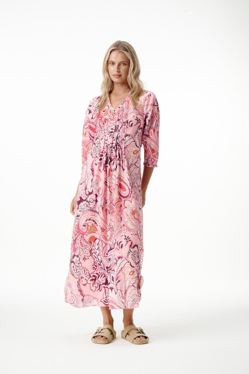 A woman modeling a pink paisley maxi dress with three-quarter sleeves and a gathered waist, paired with tan slide sandals, against a plain white studio background.
