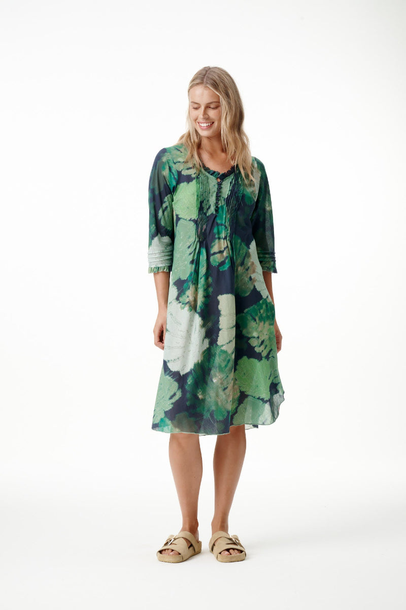 A woman modeling a knee-length, three-quarter sleeve tunic dress with a green, blue, and white botanical leaf pattern, paired with tan slide sandals, against a plain white studio background.
