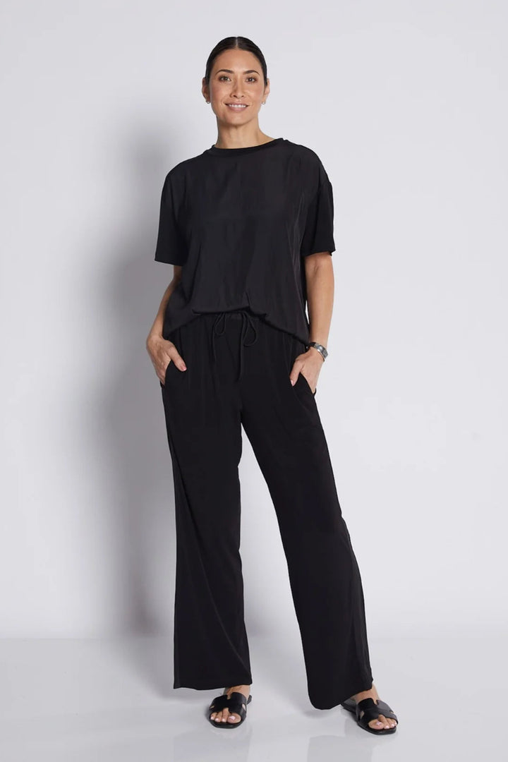 A woman modeling a black short-sleeve top and matching black wide-leg pants, standing against a light gray studio background.