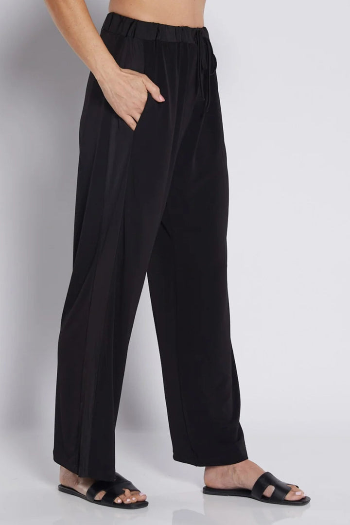 Black wide-leg trousers with an elastic waistband and side pockets, shown on a model wearing black slide sandals against a light gray studio background.