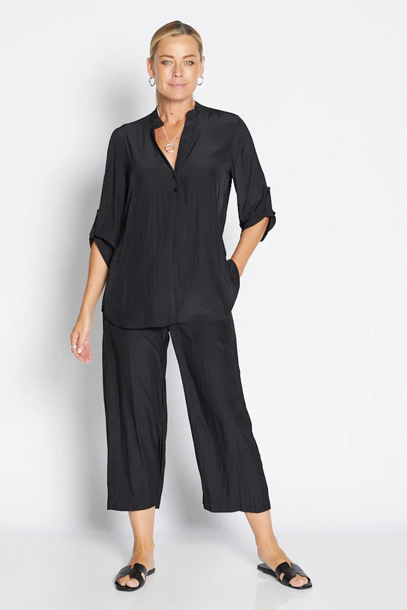 A woman modeling a black two-piece matching set consisting of a short-sleeve button-front blouse with rolled cuffs and matching wide-leg cropped pants, photographed against a clean light gray studio background.