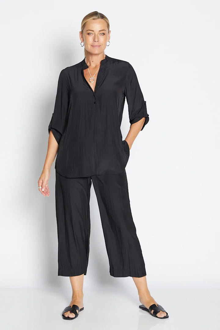 A woman modeling a black two-piece matching set consisting of a short-sleeve button-front blouse with rolled cuffs and matching wide-leg cropped pants, photographed against a clean light gray studio background.