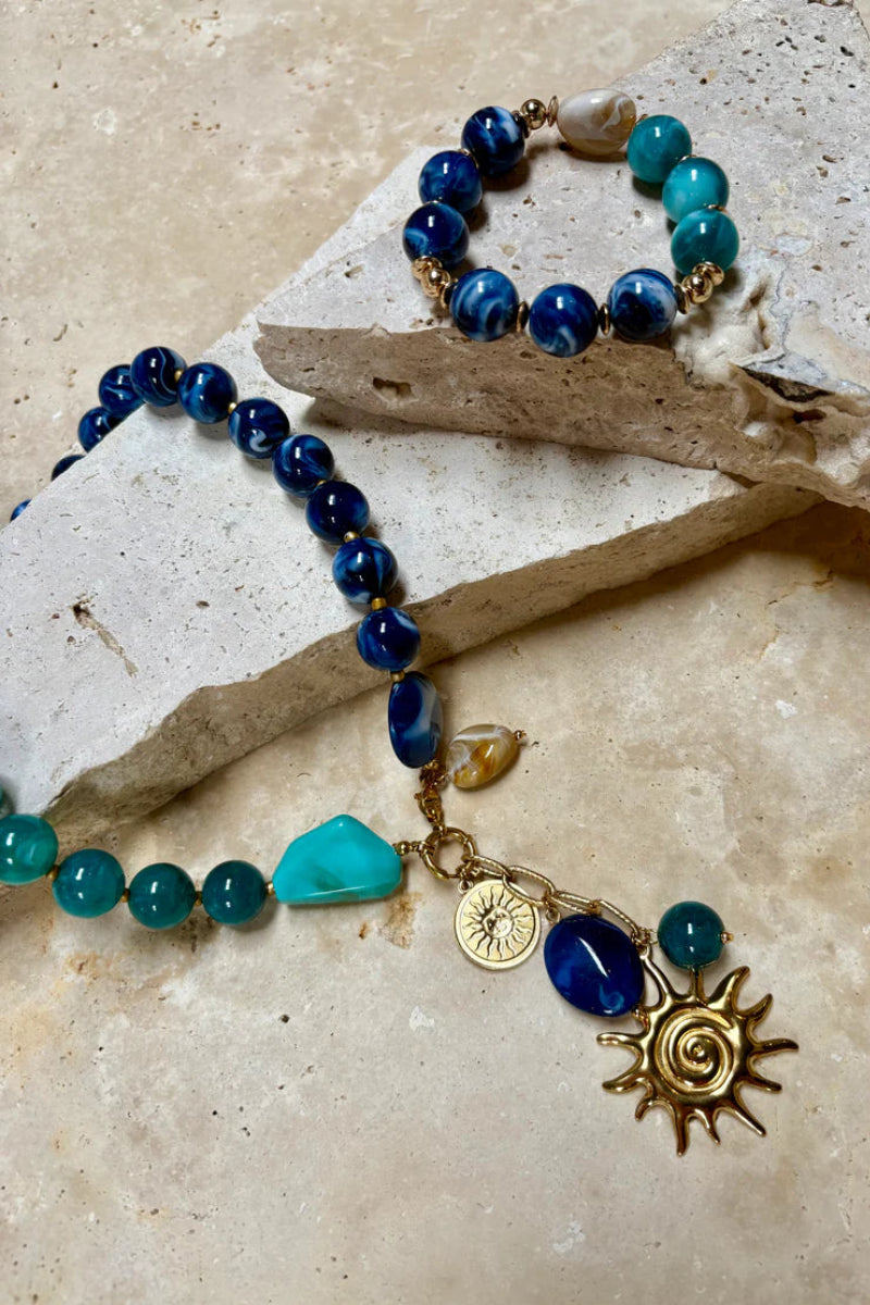 Necklace with blue and green beads and gold sun charm on a textured surface