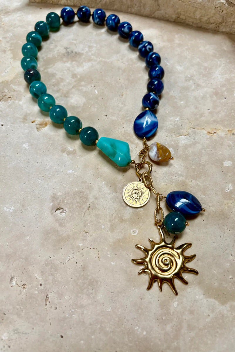 Beaded bracelet with blue and green beads and gold sun charm on a stone surface