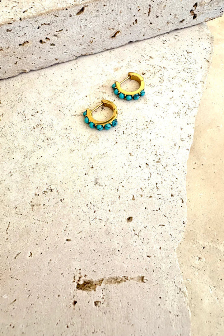 Pair of gold hoop earrings with turquoise beads on a textured beige surface.