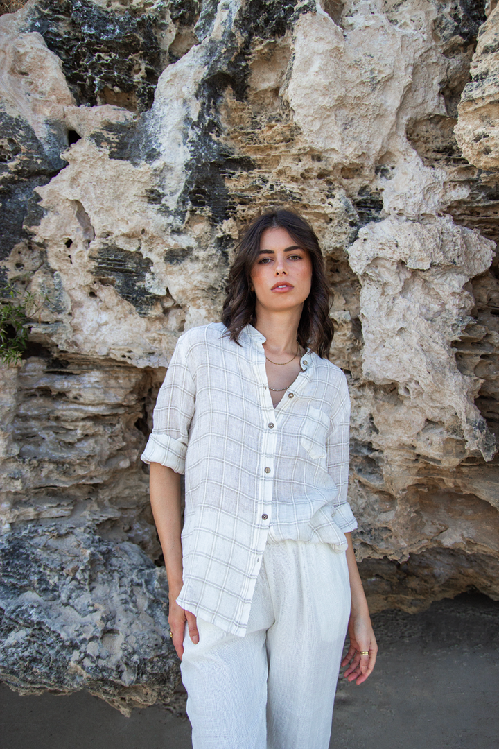 Woman in a light outfit standing against a rocky background. Shirt is checked beige and is a classic button front design with roll-tab sleeves.