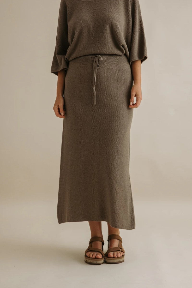 Person wearing a brown long skirt and top against a beige background