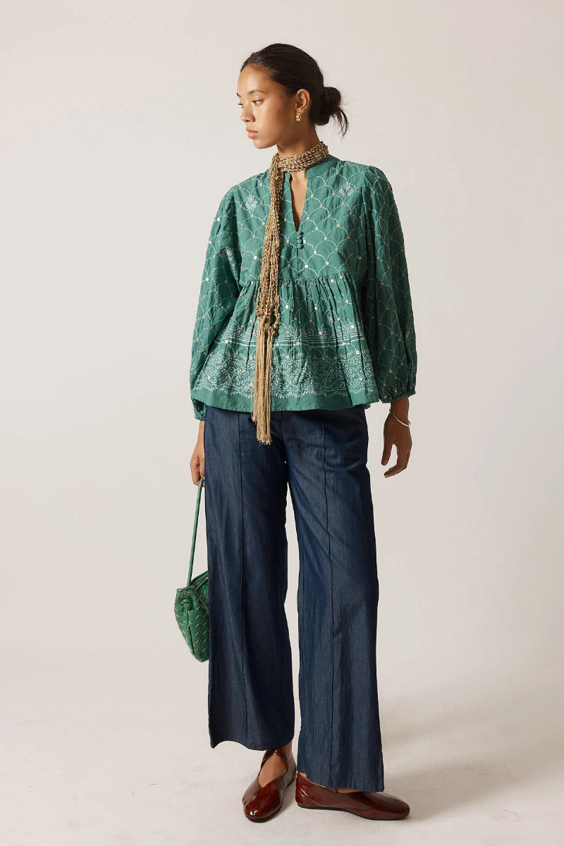 Woman wearing a green patterned top and blue pants, holding a green bag on a plain background