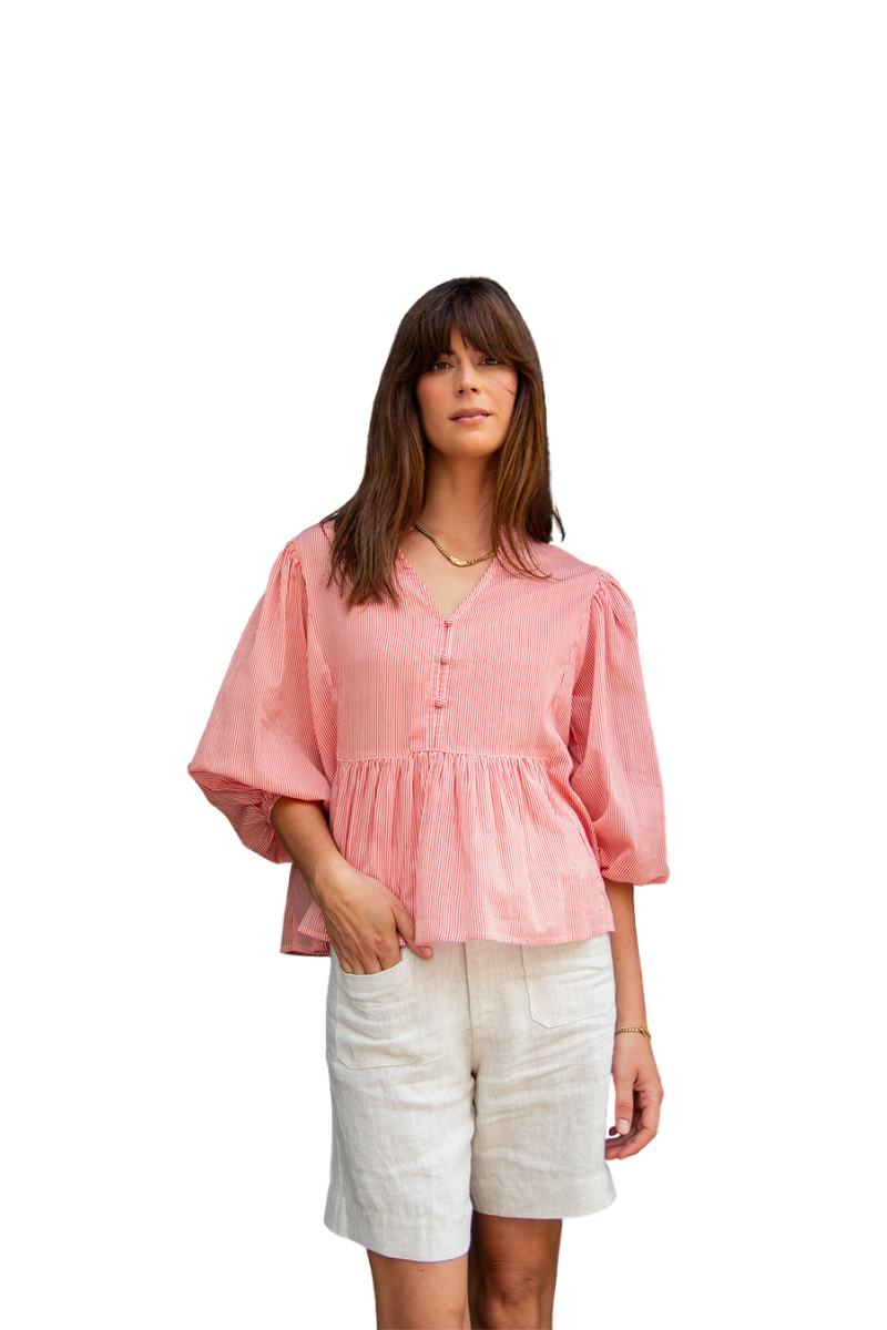 A woman modeling a light pink linen-blend peplum blouse with three-quarter puff sleeves and a buttoned V-neck, paired with off-white linen shorts, against a solid white background.