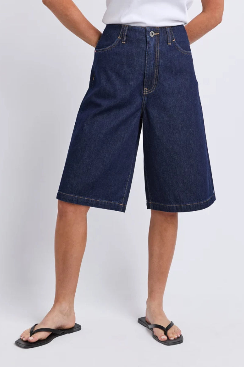 A model wearing dark blue denim shorts with a wide leg and relaxed fit, paired with a white t-shirt and black flip-flops against a light gray studio background.