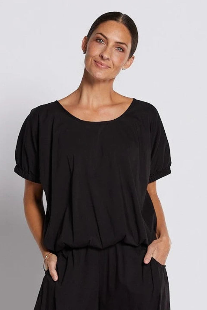 A woman modeling a black short-sleeve top with a wide scoop neckline and voluminous sleeves, paired with black pants, against a light gray studio background.