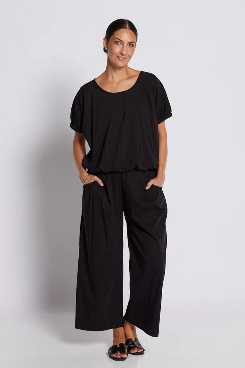 A woman modeling a black two-piece outfit consisting of a short-sleeve scoop-neck top and matching wide-leg pants, standing against a light gray studio background.