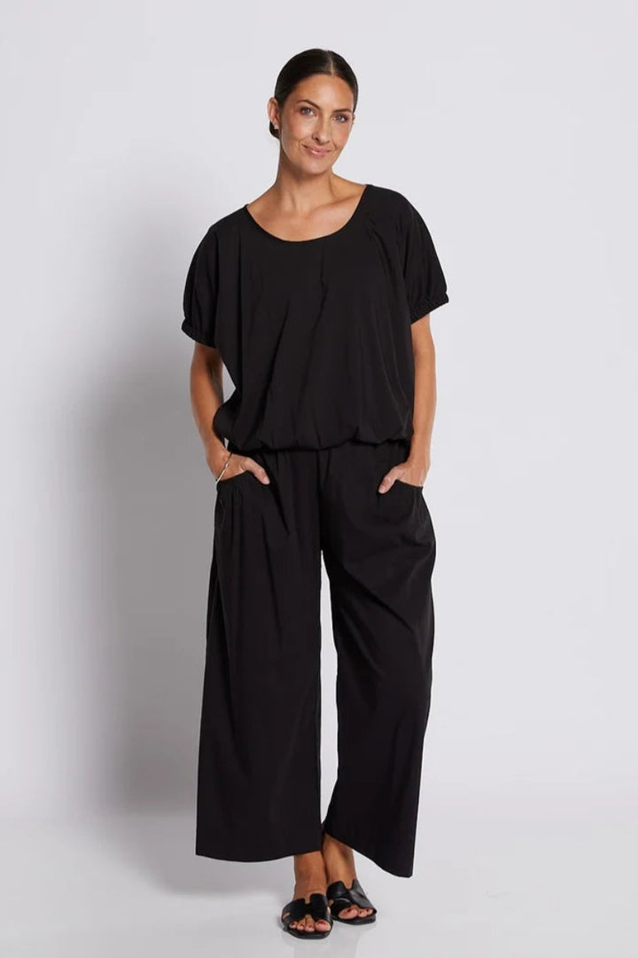 A woman modeling a black two-piece outfit consisting of a short-sleeve scoop-neck top and matching wide-leg pants, standing against a light gray studio background.