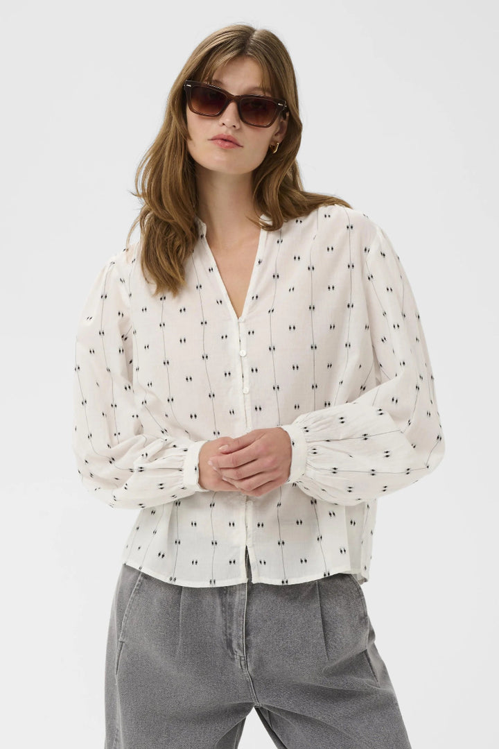 Woman wearing a white blouse with black patterns and sunglasses on a light gray background