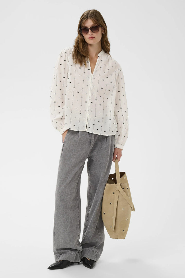Woman wearing a white blouse with a pattern, gray pants, and holding a beige bag on a white background