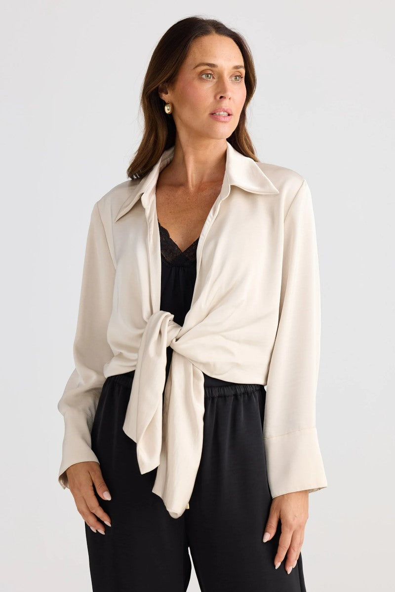 Woman wearing a beige tie-front shirt over a black top and pants on a white background
