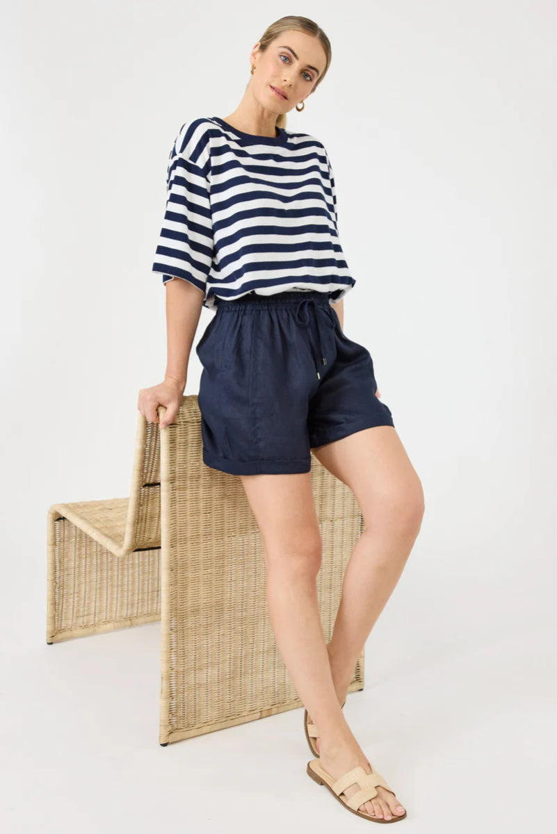 Woman wearing a navy and white striped shirt and navy shorts, standing next to a woven chair on a white background.