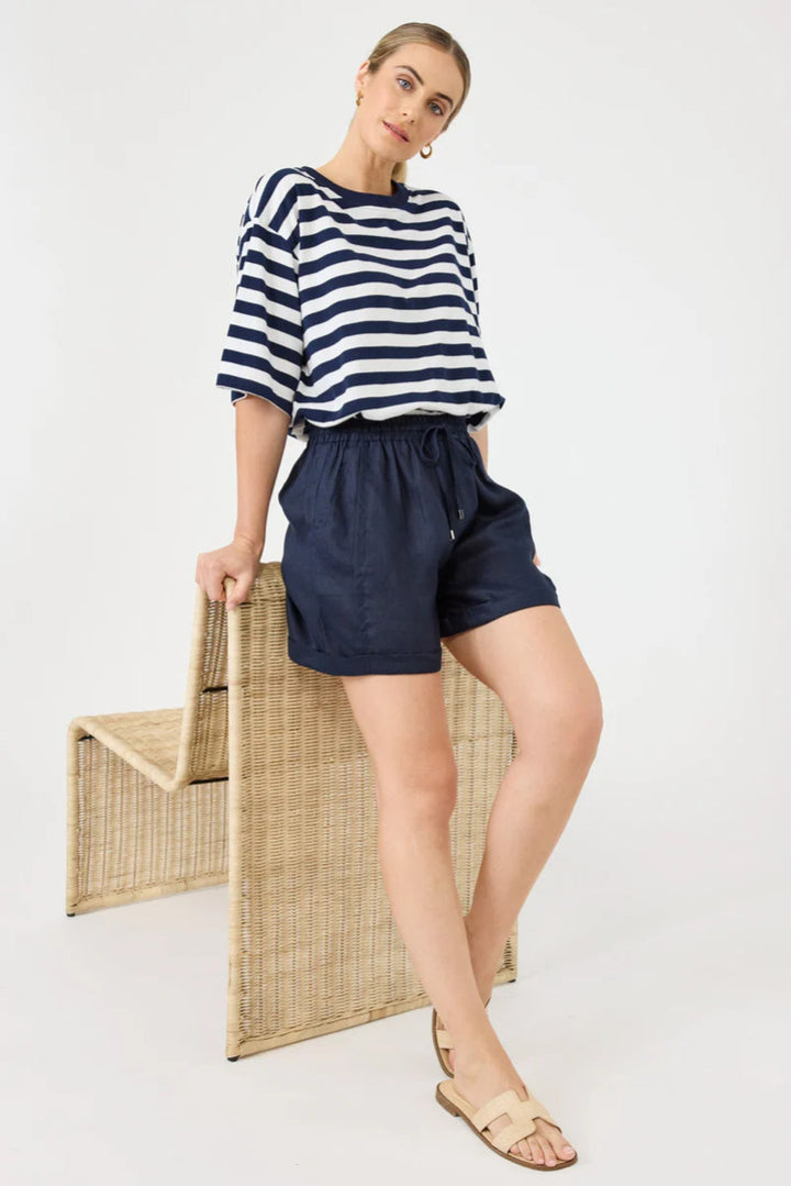 Woman wearing a navy and white striped shirt and navy shorts, standing next to a woven chair on a white background.