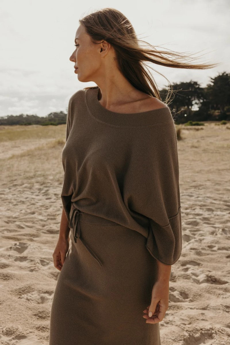 Woman wearing a brown sweater and skirt on a sandy beach.