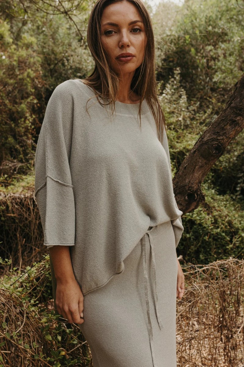 Woman wearing a light gray outfit standing in a natural setting with greenery.
