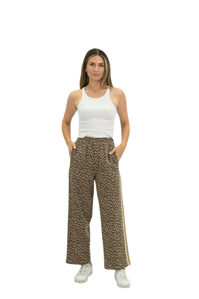 A woman modeling a pair of wide-leg brown leopard-print pants with a gold side stripe, paired with a white tank top and white sneakers, against a plain white background.