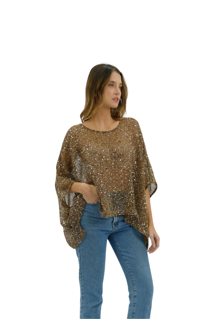 A woman modeling a sheer, metallic bronze sequined poncho top paired with blue jeans, set against a plain white background.
