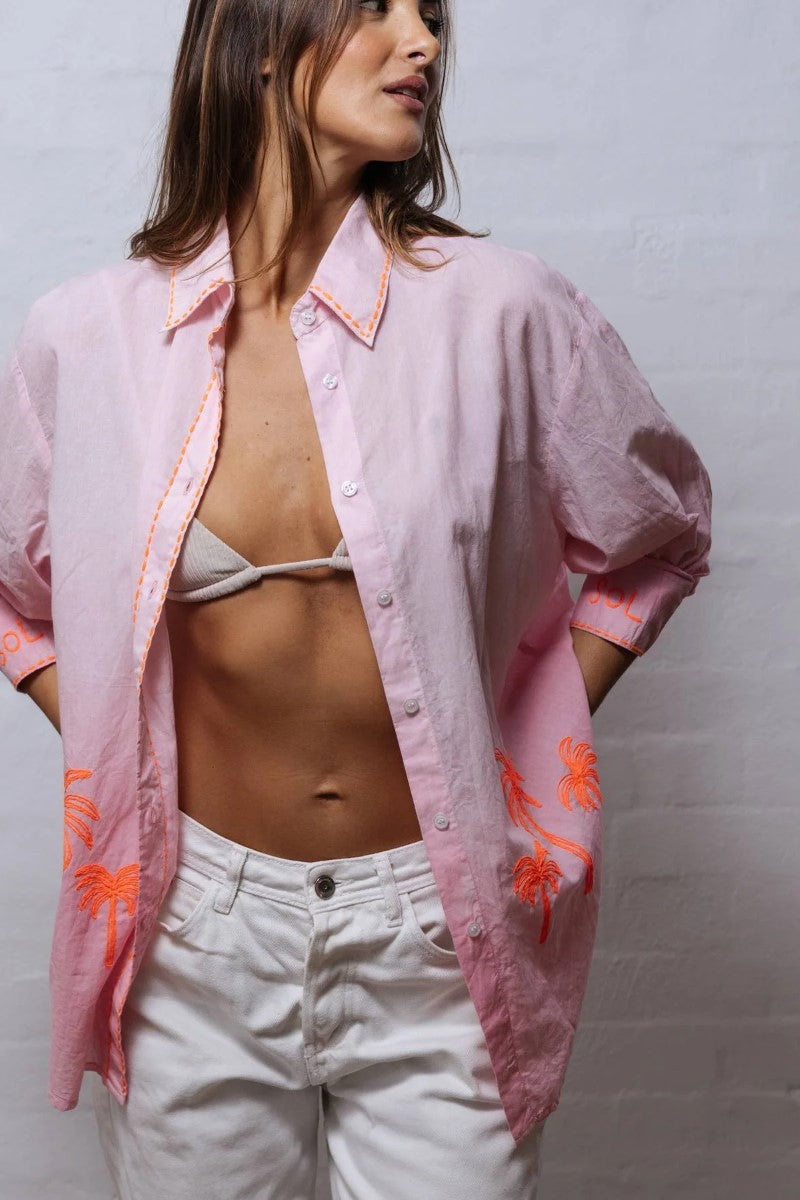 Woman wearing a pink open shirt over a bikini with white pants against a gray background