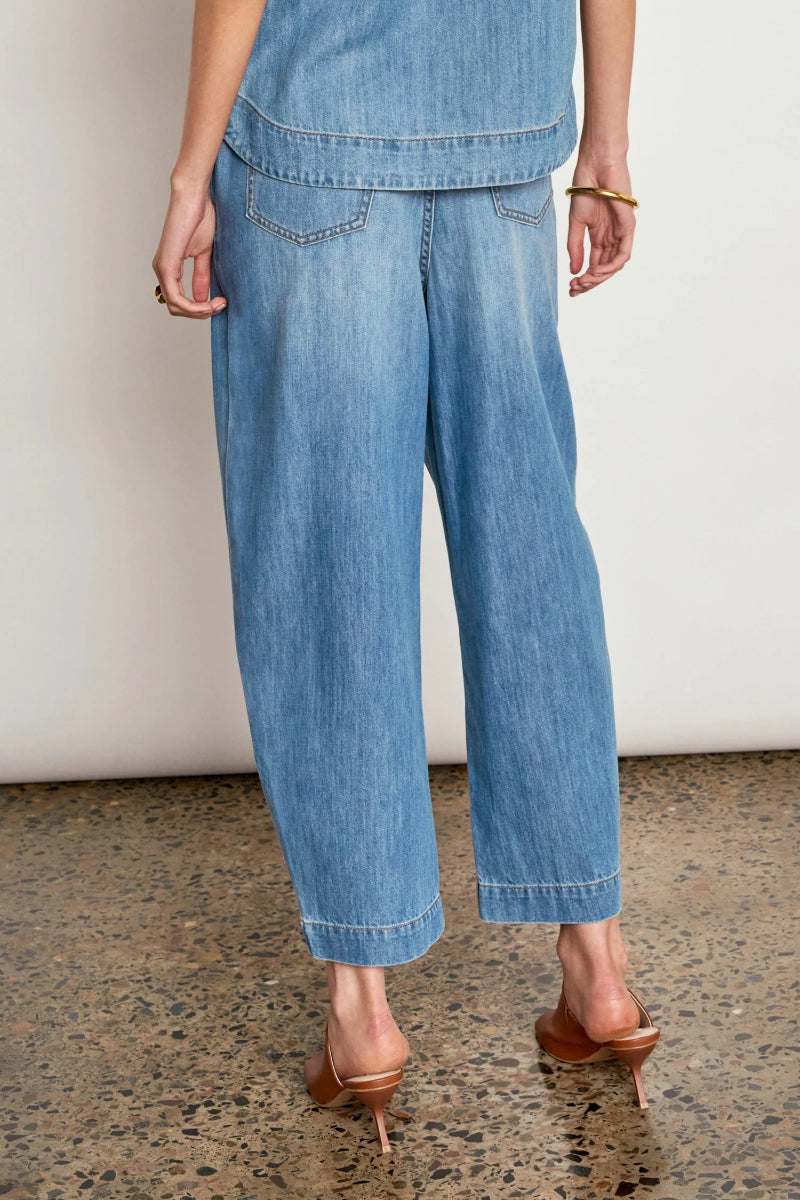 A person shown from the back wearing light blue wide-leg cropped denim jeans with rolled hems, paired with brown heeled mule sandals against a plain off-white wall and speckled floor.
