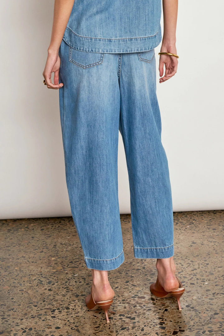 A person shown from the back wearing light blue wide-leg cropped denim jeans with rolled hems, paired with brown heeled mule sandals against a plain off-white wall and speckled floor.