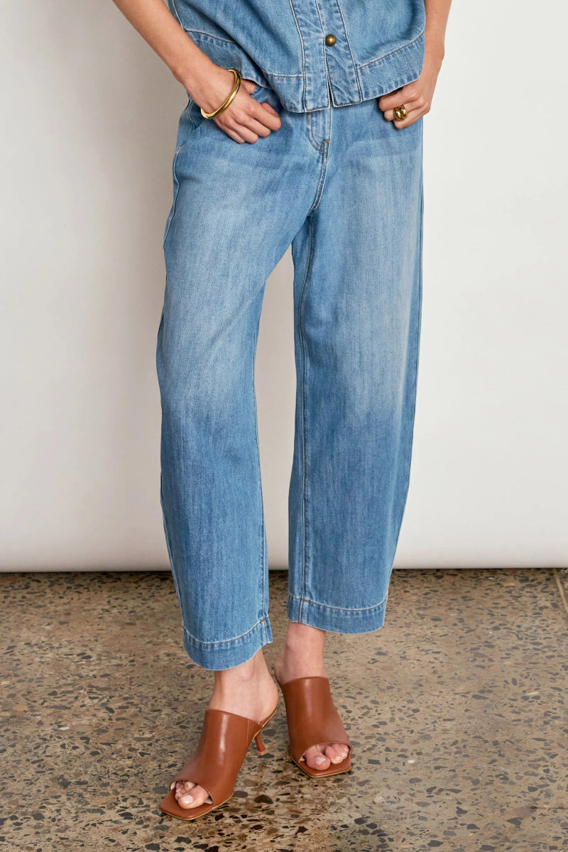 A person wearing light blue wide-leg cropped denim jeans with a matching denim shirt and brown leather mule sandals, standing against a plain off-white wall and speckled floor.
