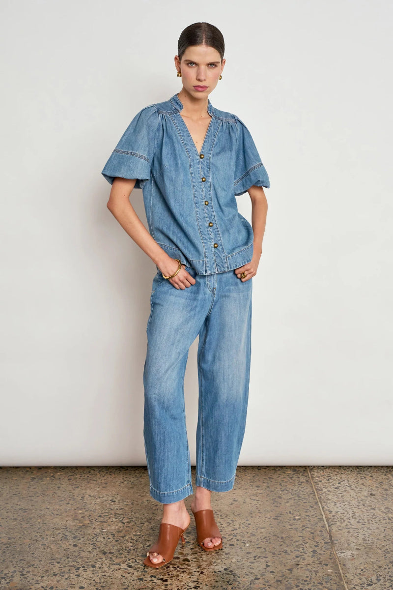 A model stands against a plain off-white wall wearing a light blue denim short-sleeve button-front top and matching wide-leg cropped jeans, paired with brown heeled sandals.
