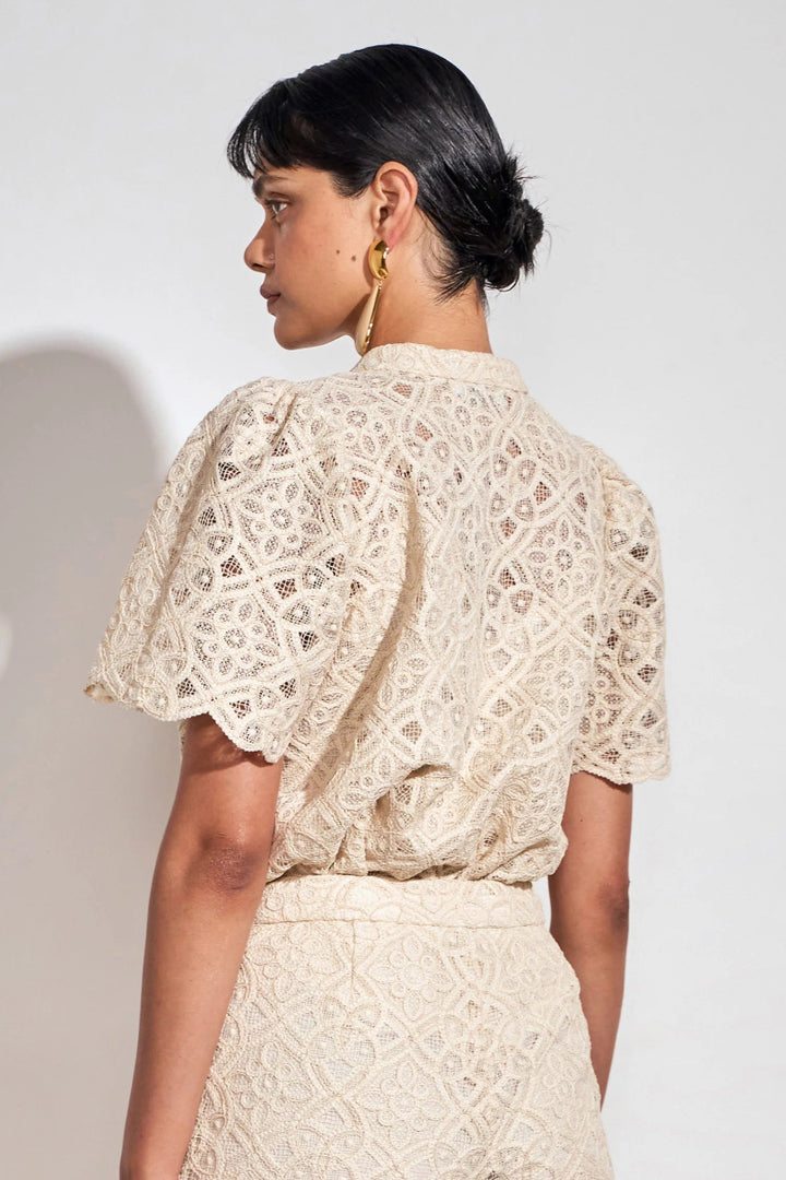 A woman shown from the back wearing a cream-colored eyelet lace top with short puff sleeves and a matching skirt against a plain light gray background.