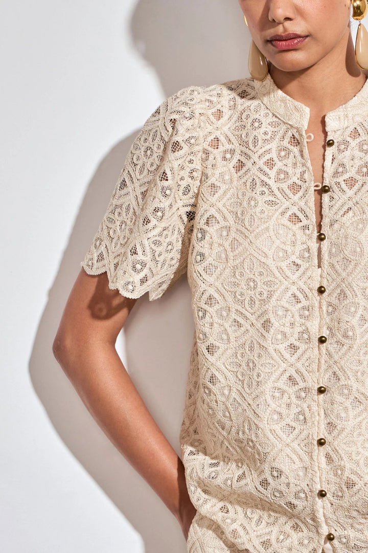 A close-up of a model wearing a cream-colored lace short-sleeve blouse with a button-down front and geometric floral lace pattern, set against a plain white background with a soft shadow.