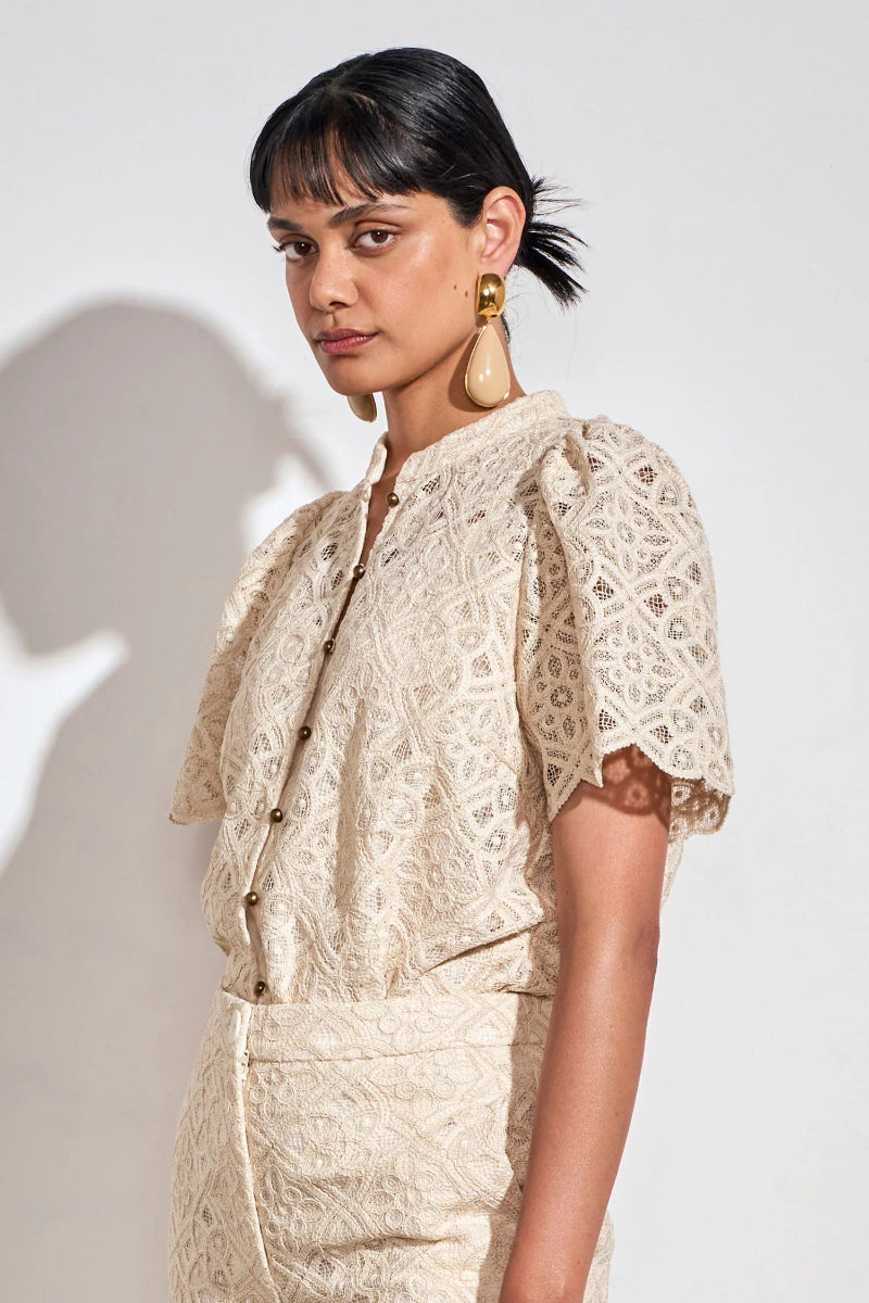 A model wearing a cream-colored lace short-sleeve top with gold buttons and matching textured skirt, styled with large gold teardrop earrings against a light gray background.