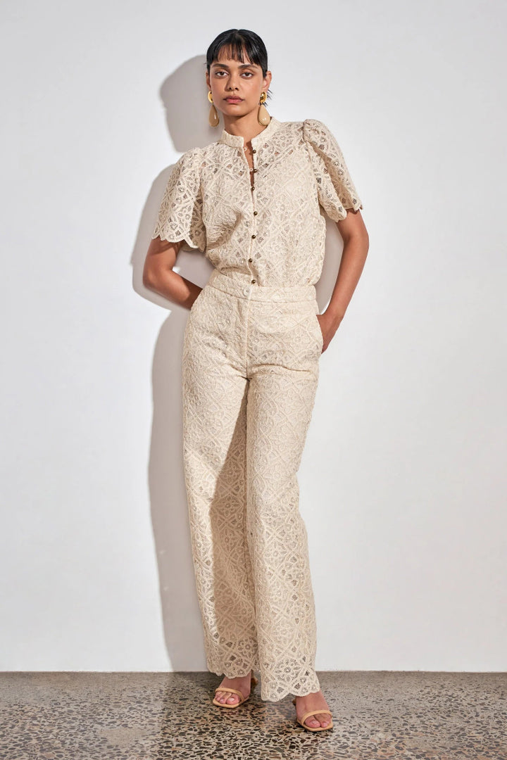 A model stands against a white wall wearing a matching cream lace top and wide-leg pants set, paired with nude flat sandals.