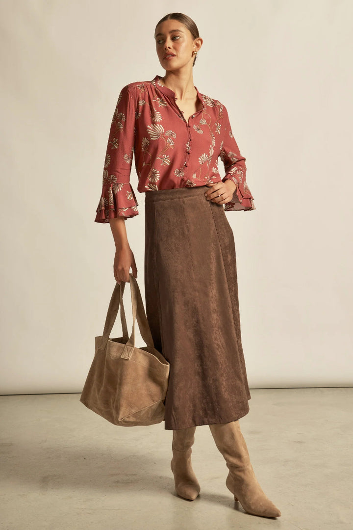 Woman wearing a red floral blouse and brown suede skirt, holding a brown bag.