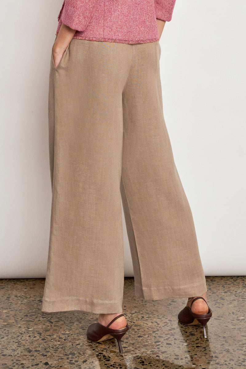 A person wearing wide-leg beige trousers, a pink textured top, and dark brown heeled sandals, standing against a plain white wall on a speckled floor.