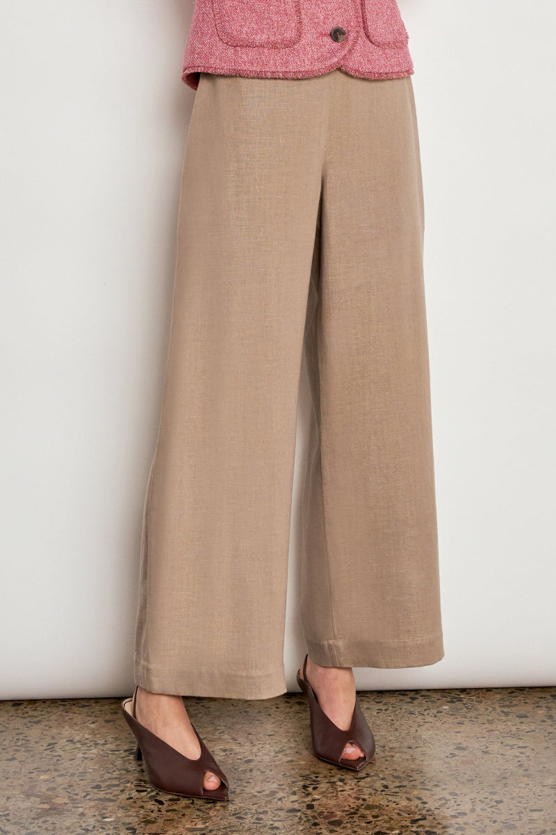 A person wearing beige wide-leg cropped trousers with a front slit, paired with a pink textured jacket and dark brown peep-toe heels, standing against a plain off-white wall.