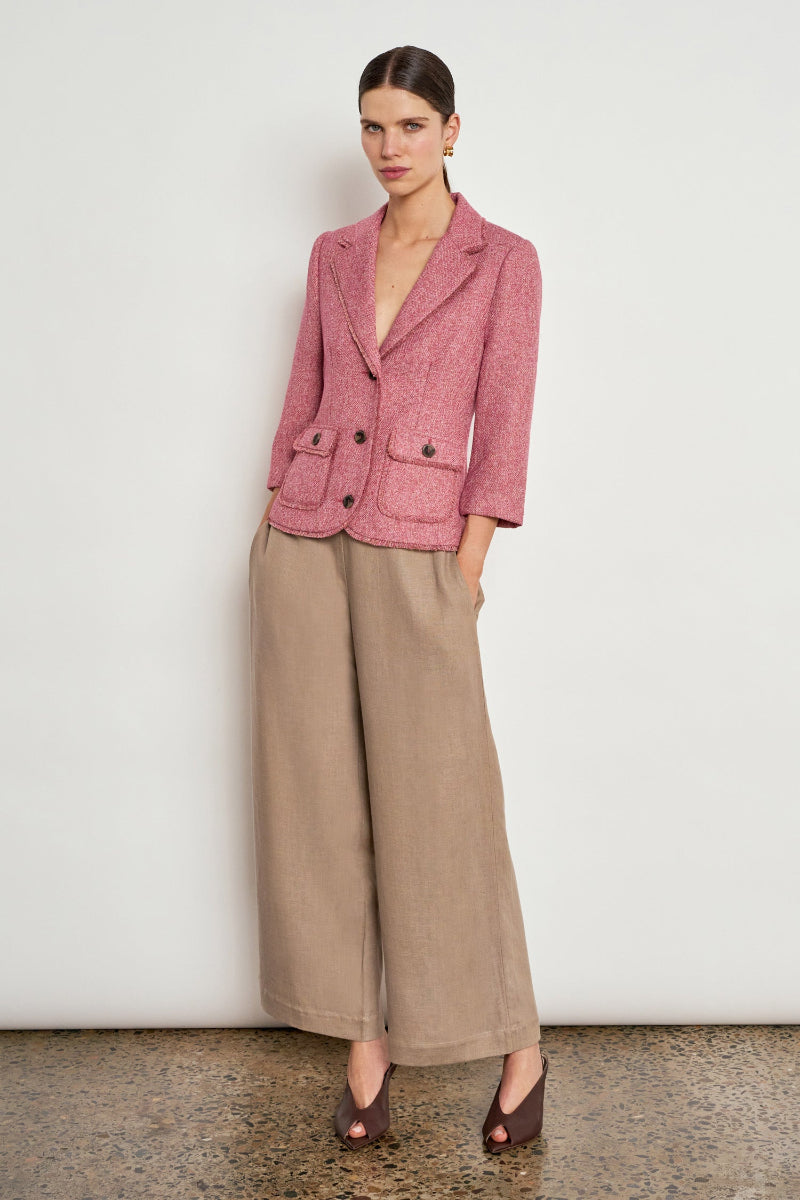 A model stands against a plain off-white wall wearing a textured dusty pink blazer and wide-leg taupe trousers, paired with dark brown pointed-toe heels.
