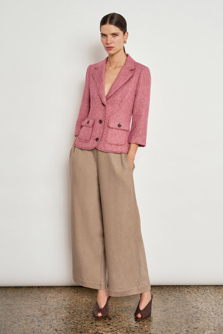 A model stands against a plain off-white wall wearing a textured dusty pink blazer and wide-leg taupe trousers, paired with dark brown pointed-toe heels.