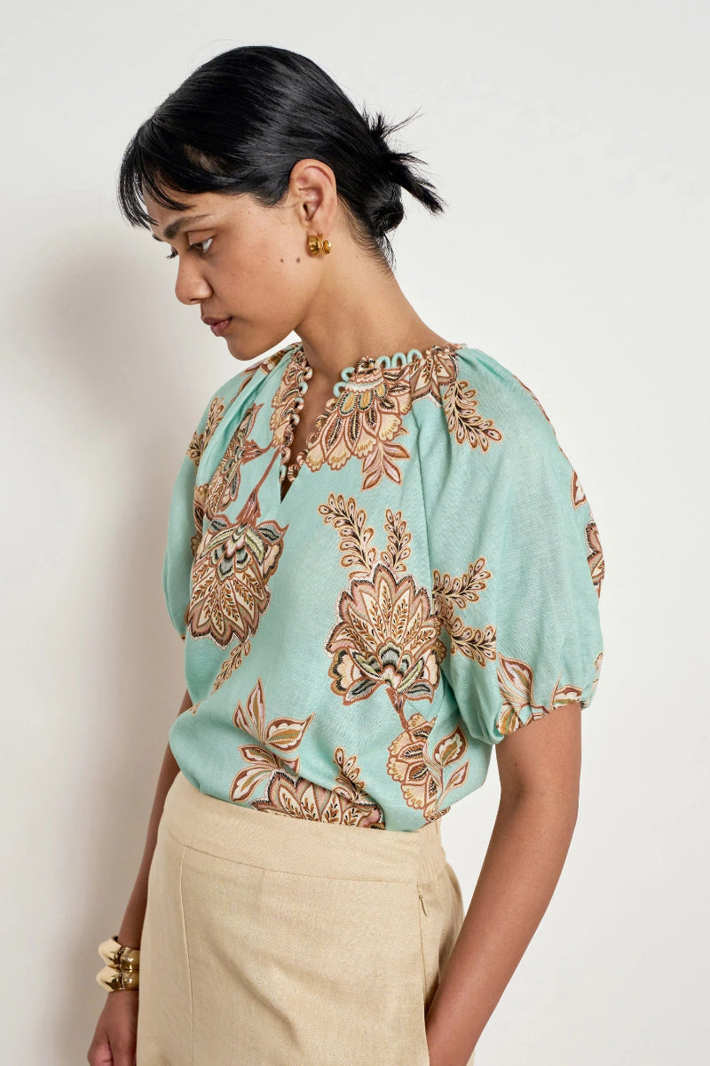 A woman wearing a light mint green short-sleeve blouse with a brown and gold floral pattern, paired with beige trousers, photographed against a plain off-white background.