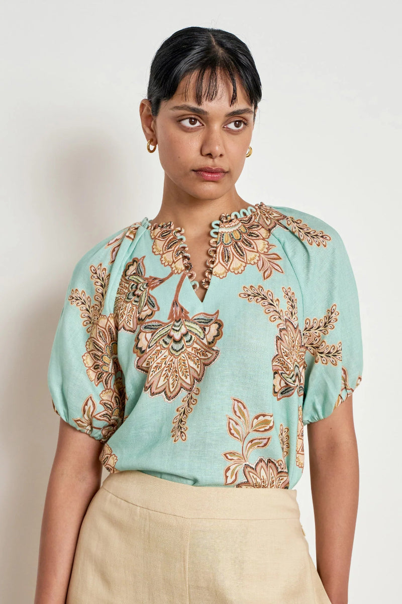 A woman wearing a light mint green blouse with brown and beige floral patterns and beige trousers, photographed against a plain off-white background.
