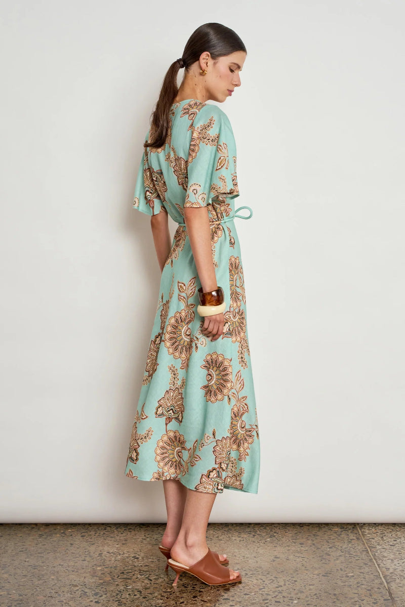 A woman modeling a light aqua midi dress with a brown and beige floral pattern, featuring flutter sleeves and a tie waist, standing against a white wall.