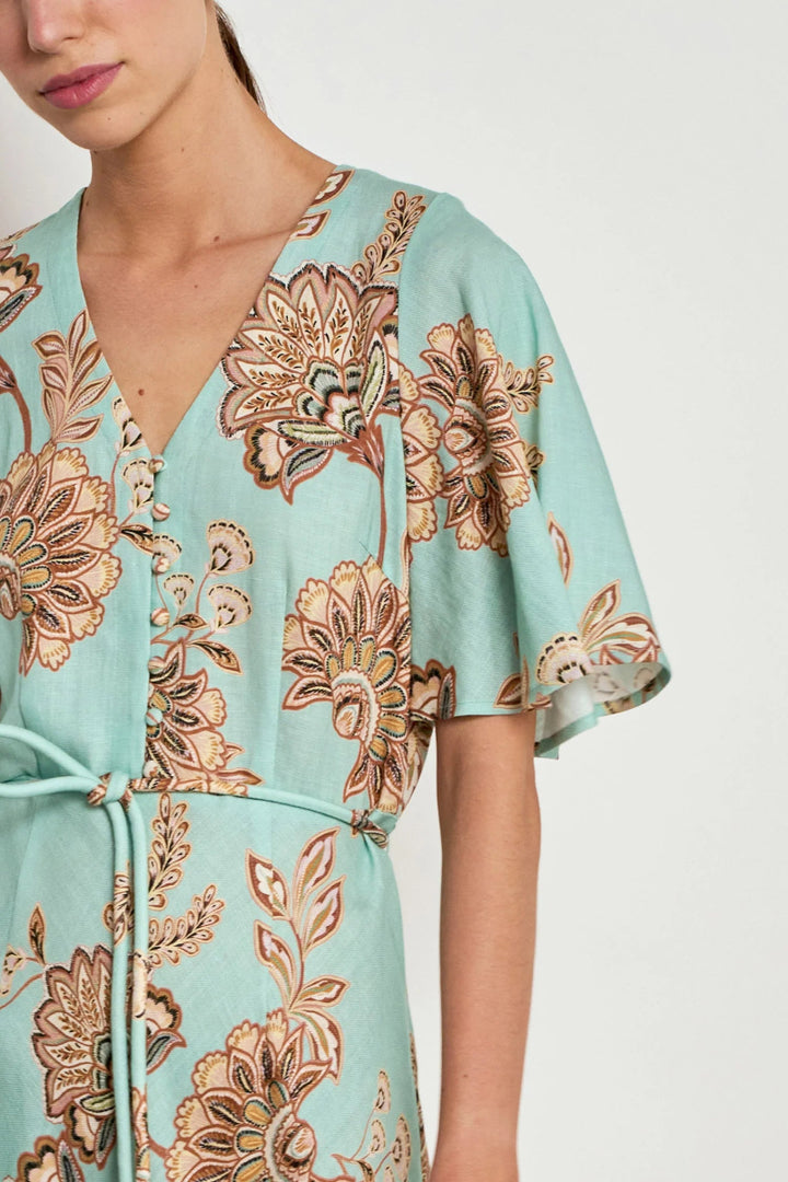 A close-up of a model wearing a light mint green wrap dress with a brown and beige floral pattern, featuring a V-neckline and short flutter sleeves.