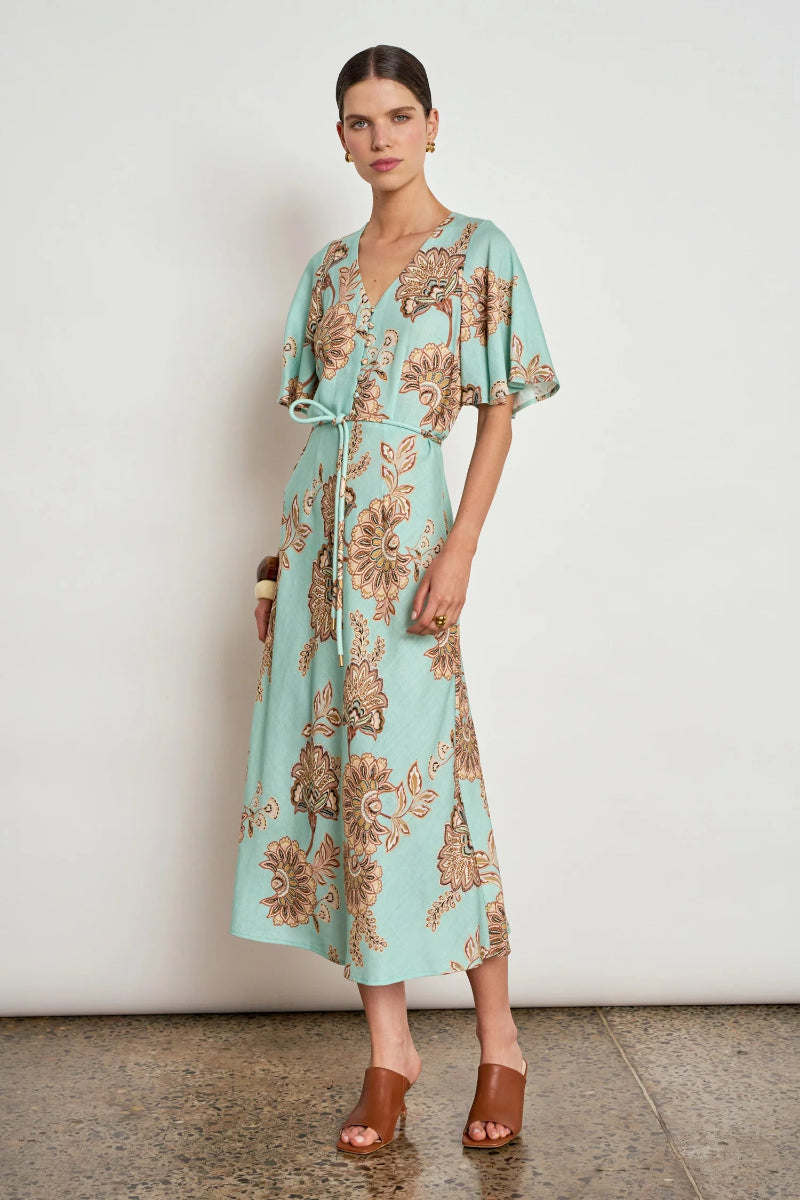 A woman models a light aqua midi wrap dress with a brown floral pattern, featuring flutter sleeves and a tie waist, paired with brown leather sandals against a neutral studio background.