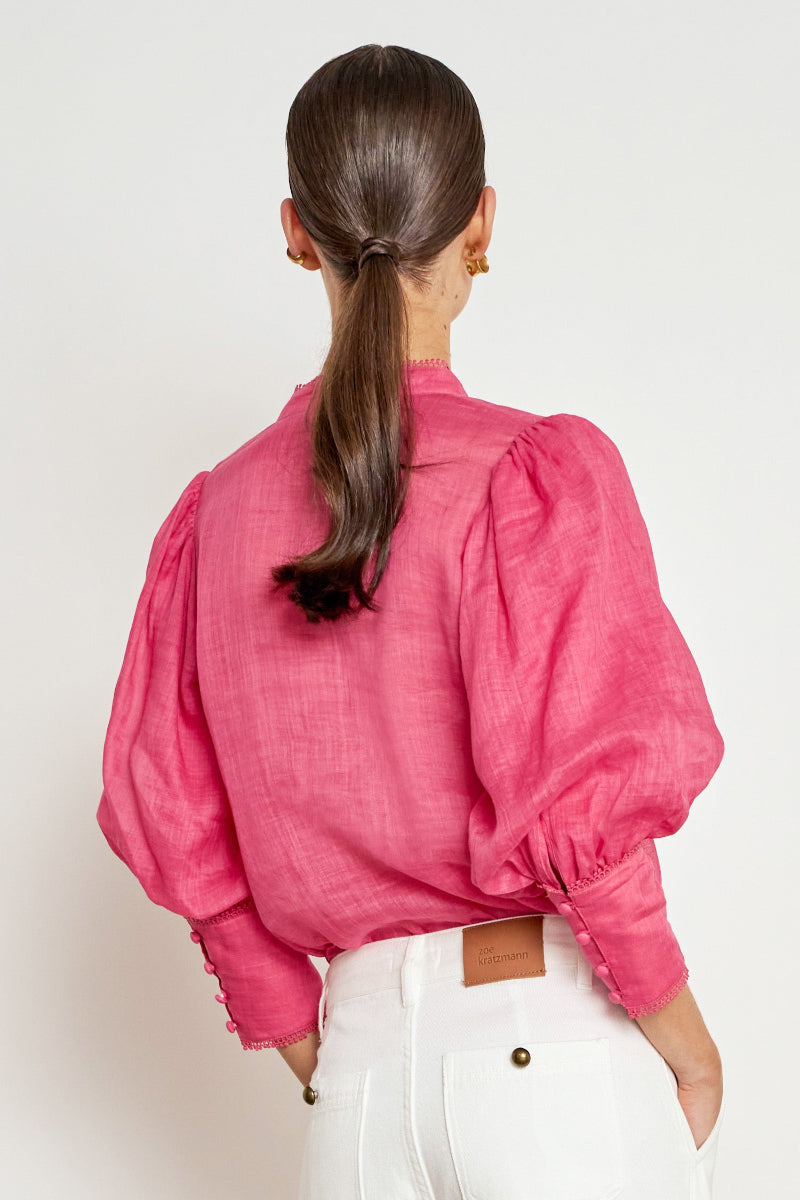A woman viewed from the back wearing a bright pink long-sleeve blouse with voluminous puffed sleeves and white pants, against a plain light background.