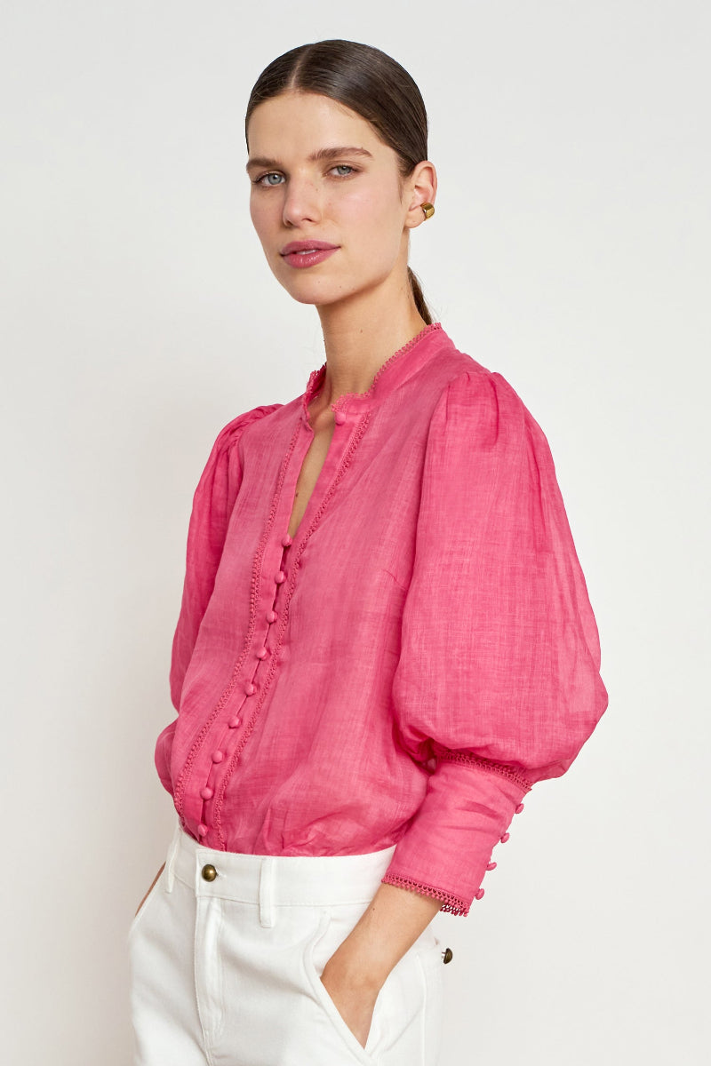 A woman wearing a bright pink long-sleeve blouse with puffed balloon sleeves and a buttoned collar, paired with white pants, photographed against a plain light gray studio background.