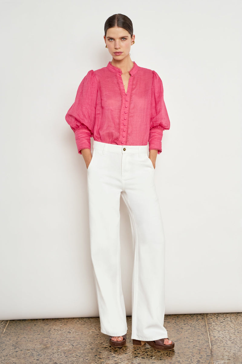 A model stands against a plain off-white wall wearing a bright pink long-sleeve button-up blouse with puffed shoulders and white wide-leg trousers.