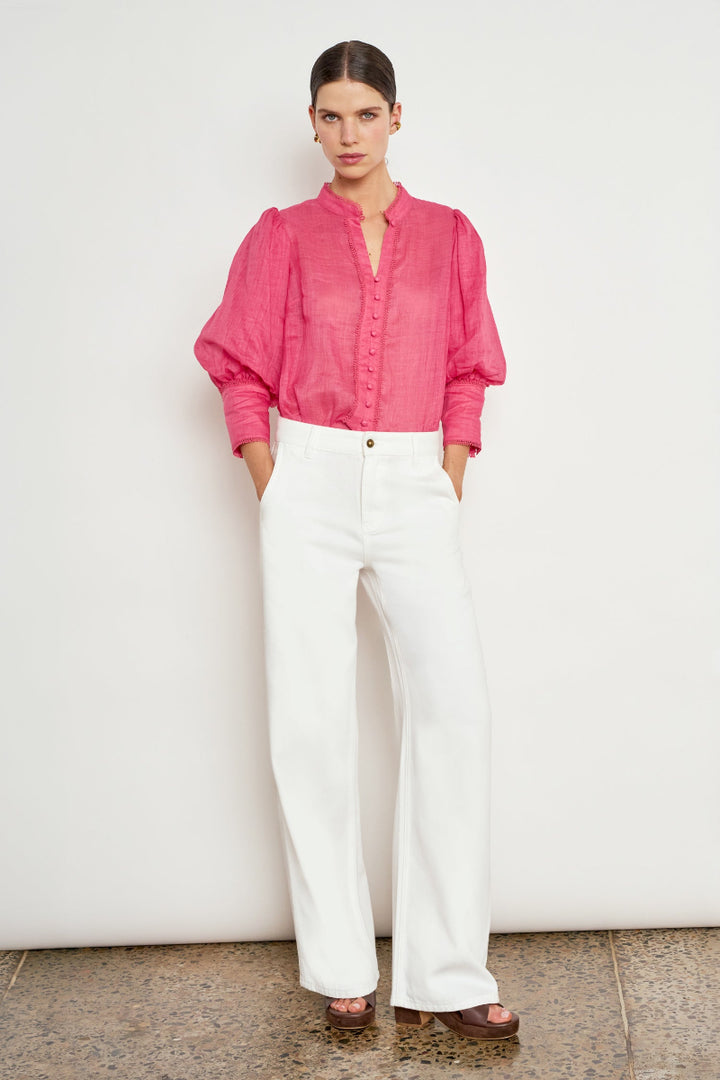 A model stands against a plain off-white wall wearing a bright pink long-sleeve button-up blouse with puffed shoulders and white wide-leg trousers.