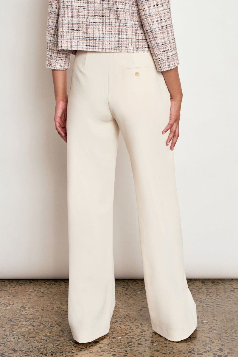 A person shown from the back wearing cream wide-leg trousers and a beige plaid cropped shirt, standing against a plain off-white wall on a speckled floor.